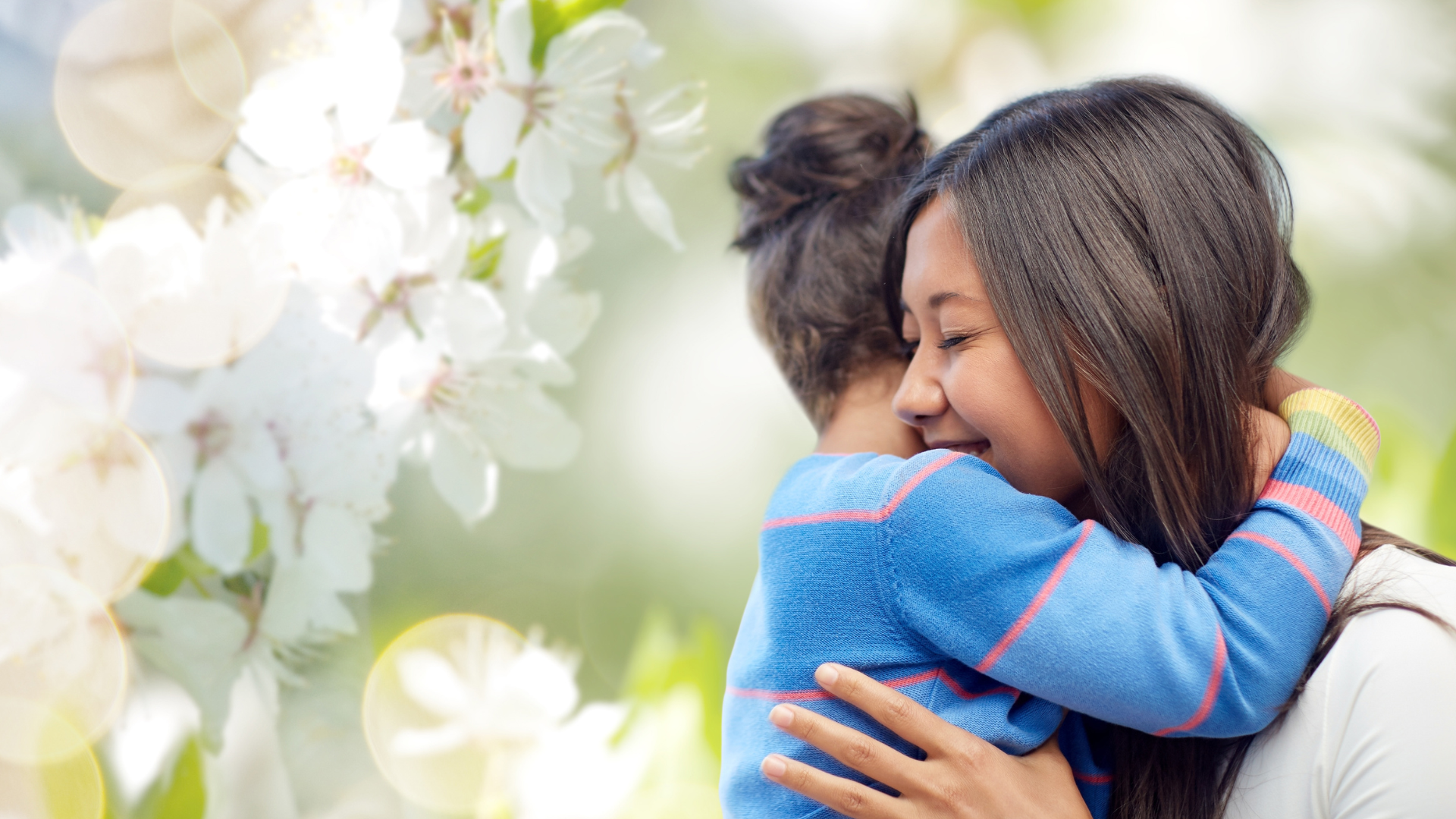 A photo of a mother smiling while her child hugs her around her shoulders