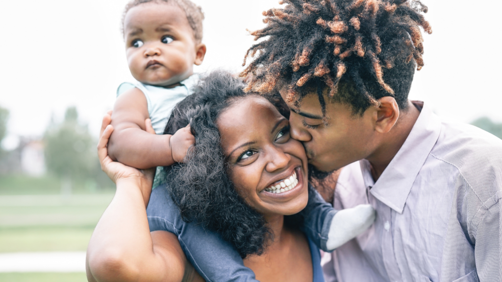 A man kissing a woman’s cheek while she holds a baby on her shoulders