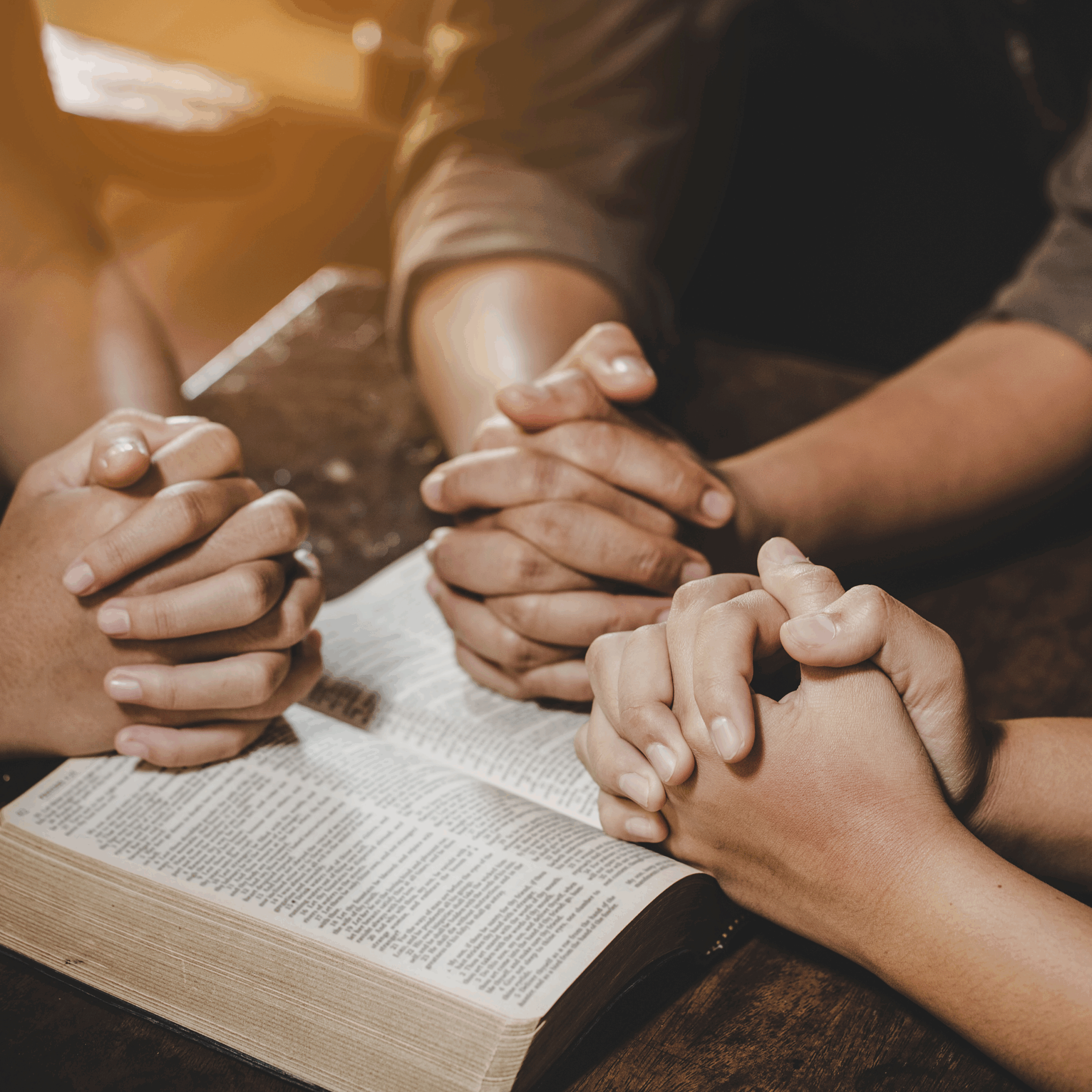 Three peoples hands folded in prayer over a Bible